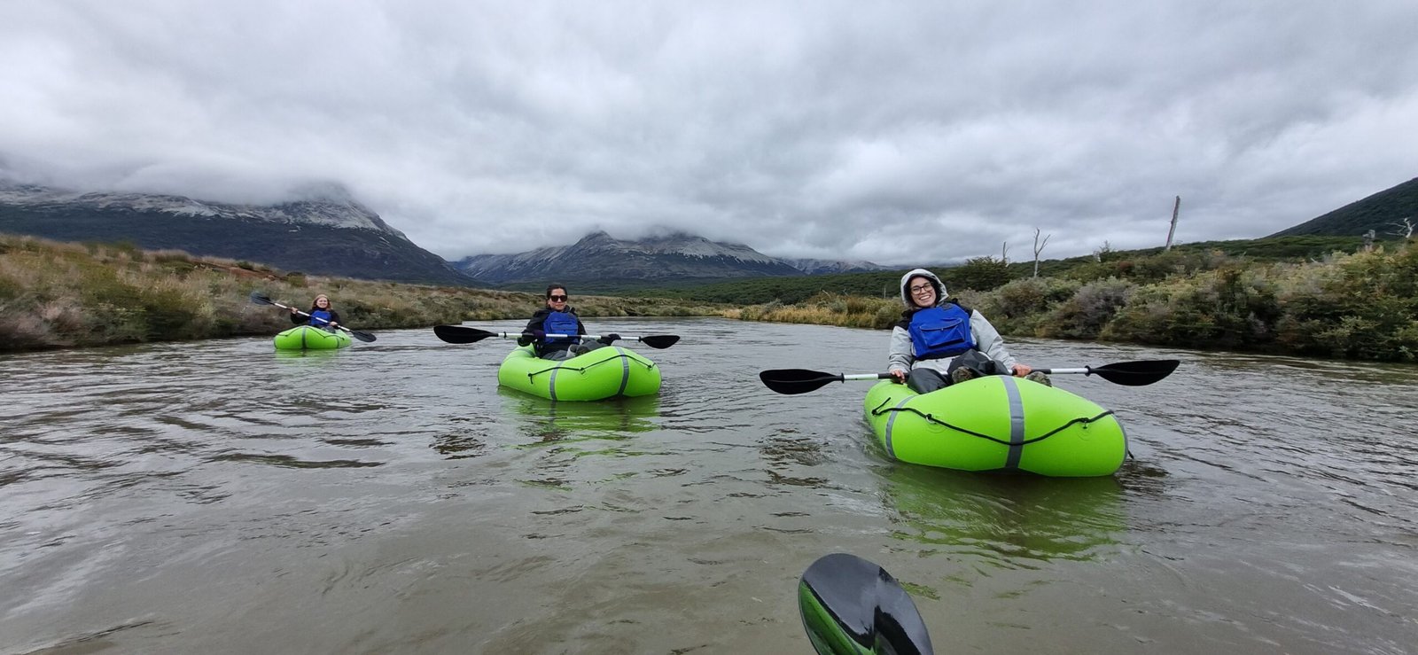 Tres turistas sonrientes remando en packrafts de color verde brillante por un río en Ushuaia, con las montañas de la Cordillera de los Andes cubiertas por nubes bajas al fondo. Excursión de aventura con XPLOR Turismo.