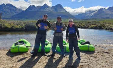 Tres turistas sonrientes equipados con chalecos salvavidas azules, posando junto a tres packrafts verdes en la orilla de un río en Ushuaia, con los picos nevados de los Andes Fueguinos de fondo. Excursión de aventura con XPLOR Turismo.