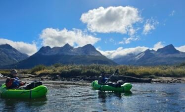 Dos personas remando en packrafts verdes sobre las aguas cristalinas del Río Olivia en Ushuaia, con los picos imponentes de la Cordillera de los Andes de fondo bajo un cielo azul con nubes. Excursión guiada con XPLOR Turismo