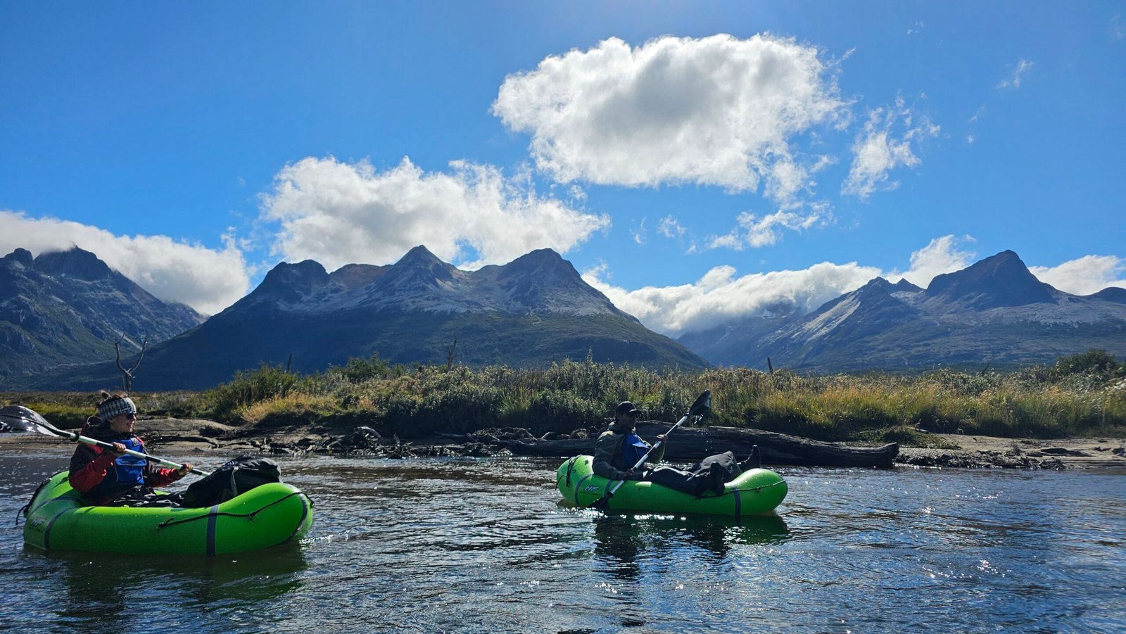 Dos personas remando en packrafts verdes sobre las aguas cristalinas del Río Olivia en Ushuaia, con los picos imponentes de la Cordillera de los Andes de fondo bajo un cielo azul con nubes. Excursión guiada con XPLOR Turismo